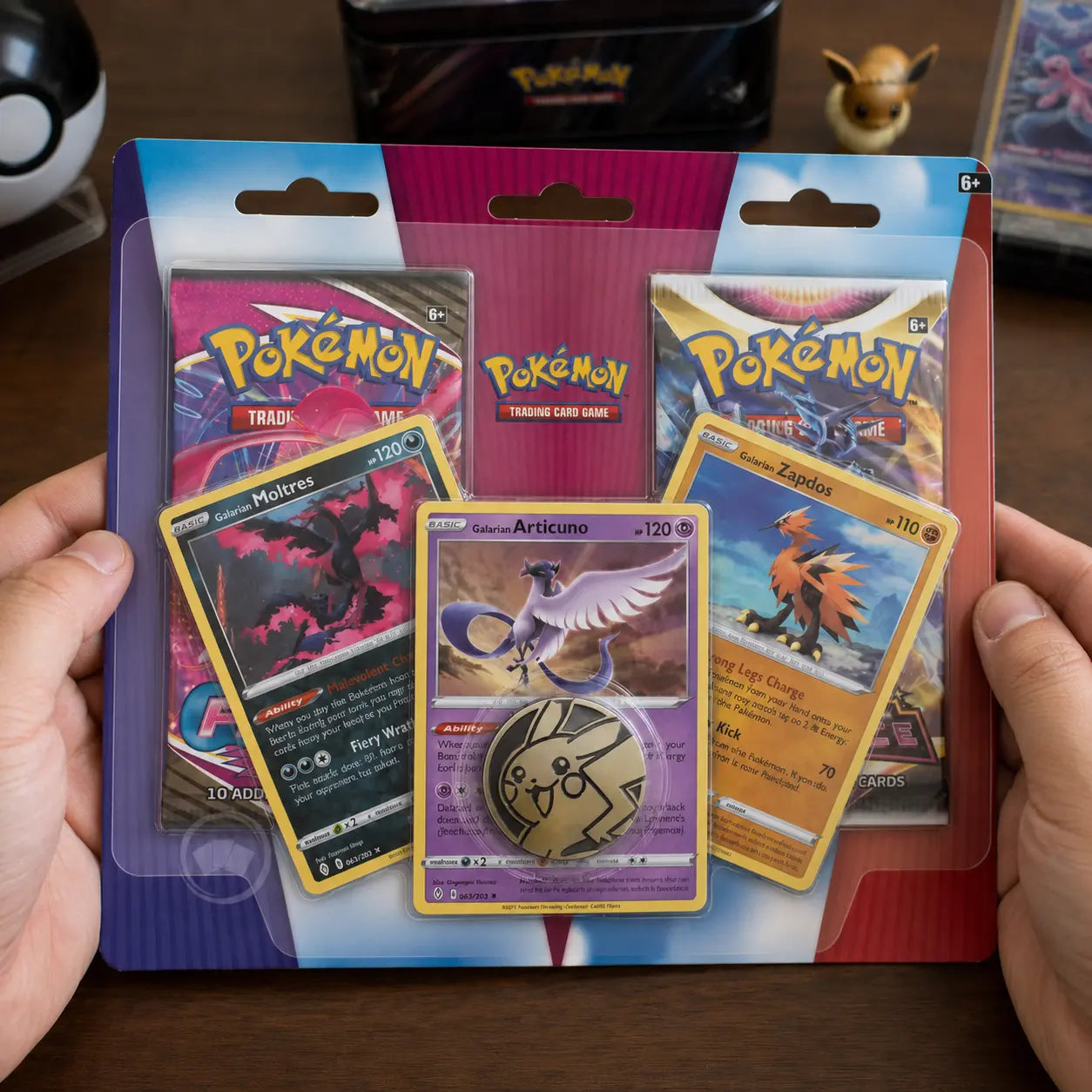 Hände halten Pokemon Galarian Birds Trio 2-Pack Blister auf Holztisch mit Evoli-Figur im Hintergrund